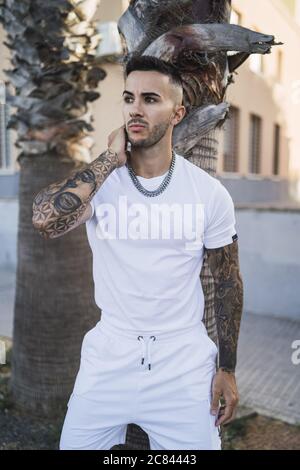 Vertical shot of a young European tattooed hip male in white shorts and shirt and a metal chain Stock Photo