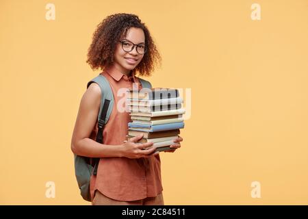 Portrait of smiling smart Afro-American student girl with satchel on back carrying stack of books against yellow background Stock Photo