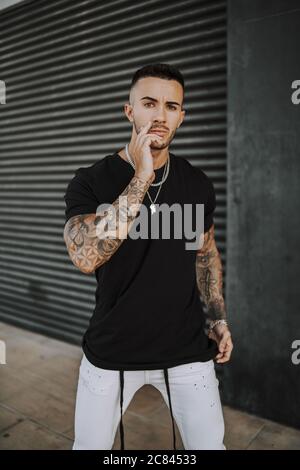 Vertical shot of a young tattooed hip male touching his face while posing  in front of a garage Stock Photo