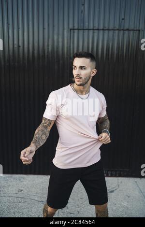 Young European tattooed hip male touching his shirt standing in front of a garage Stock Photo