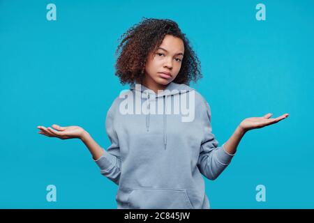 Portrait of distraught Afro-American girl in hoodie shrugging shoulders against blue background, confusion concept Stock Photo