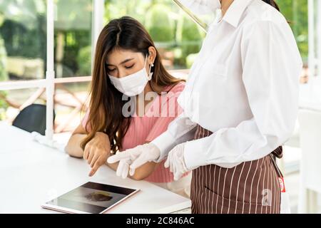 Asian waitress wear facemask and face shield using tablet to show restaurant electronic menu and recommend to customer. New normal technology and life Stock Photo