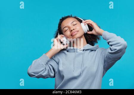 Calm relaxed Afro-American girl in wireless headphones keeping eyes closed while listening to music against blue background Stock Photo