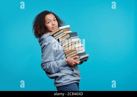 Portrait of content curly-haired black student girl carrying big stack of books against blue background, preparing for exam concept Stock Photo