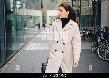 Young businesswoman in coat confidently walking around city street with suitcase Stock Photo
