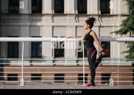 Athlete woman preparing for run on city street. African american girl in sportswear doing warm up of leg muscles in stadium in morning Stock Photo