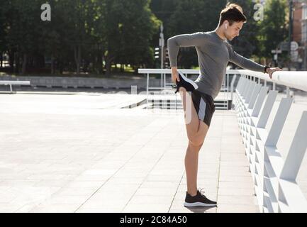 Useful leisure and preparation for marathon. Guy stretches leg near railing in park Stock Photo