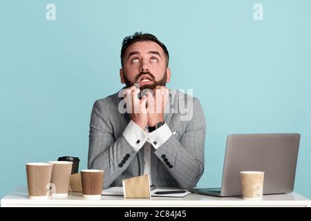 Office employee surrounded by empty coffee cups suffering from work overload at desk, blue background Stock Photo