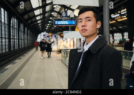 Young confident Asian businessman in wireless earphones waiting train at station Stock Photo