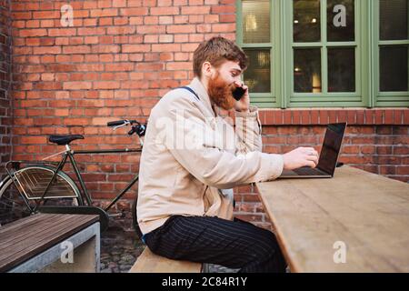 Young bearded man happily talking on cellphone working on laptop in cafe on street Stock Photo