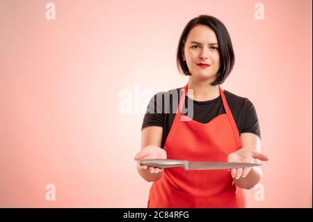 Woman employed at supermarket with red apron and black t-shirt offer a knife isolated on red background Stock Photo