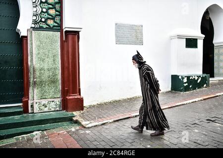 Morocco, Tangier: Scene from everyday life with inhabitants in the medina. Elderly man in striped bournous walking alone in a lane, with hands behind Stock Photo