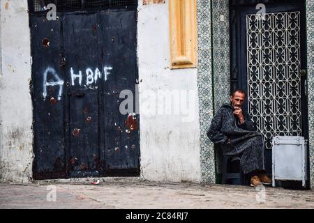Morocco, Tangier: scene from everyday life in the medina. Old man in grey bournous sitting on a chair on his doorstep Stock Photo