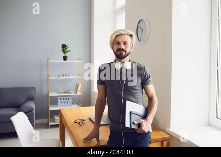 A young student guy with headphones and books in his hands stands looking at the camera in the living room. Stock Photo