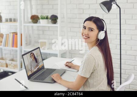 Smiling young girl in headphones communicating with her boss or teacher online and taking notes in copybook, indoors Stock Photo