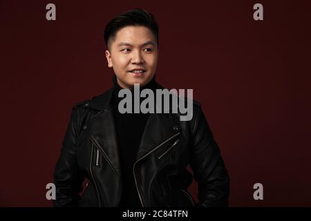 Image of joyful asian man in leather jacket smiling and looking aside isolated over burgundy background Stock Photo