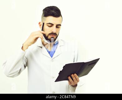 Doctor with beard reads history of present illness in notebook. Treatment and ambulance service concept. Dentist in surgical mask isolated on white background. Man with thinking face in white coat Stock Photo