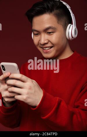 Image of joyful young asian man smiling while using headphones and cellphone isolated over burgundy background Stock Photo