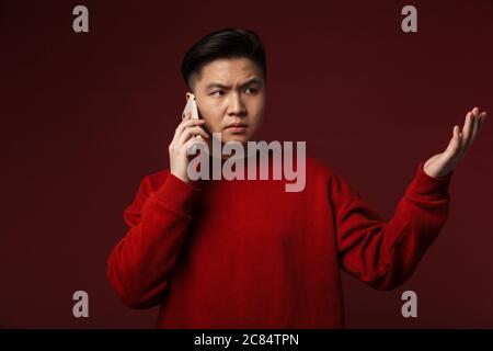 Image of displeased young asian man talking on mobile phone isolated over burgundy background Stock Photo