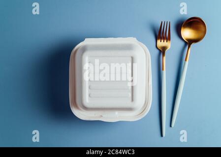 Healthy food concept: white burguer packaging closed with golden fork and spoon,  in cardboard lunch box to take to the office on blue surface Stock Photo