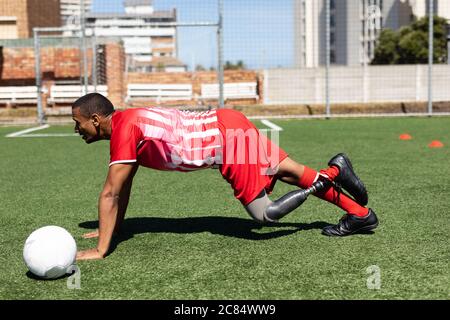 Mixed race male football player with prosthetic leg wearing a team strip training at a sports field in the sun, warming up doing push ups with ball ne Stock Photo