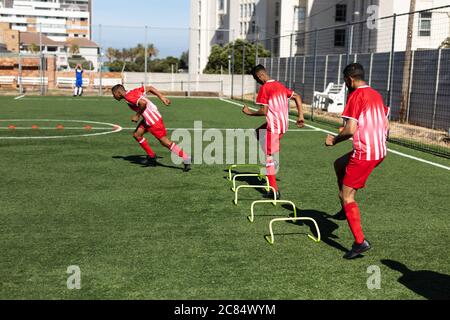 Multi ethnic team of male five a side football players wearing a team strip training at a sports field in the sun, warming up running jumping over hur Stock Photo