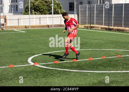 Mixed race male football player wearing a team strip training at a sports field in the sun, warming up jumping over cones. Stock Photo