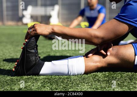 Mixed race male football player wearing a team strip training at a sports field in the sun, warming up stretching his legs. Stock Photo