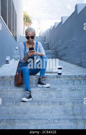 Mixed race alternative woman with short blonde hair out and about in the city on a sunny day, wearing sunglasses and denim dungarees, sitting on steps Stock Photo