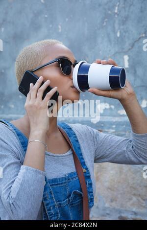 Mixed race alternative woman with short blonde hair out and about in the city on a sunny day, using smartphone, wearing sunglasses and drinking a take Stock Photo