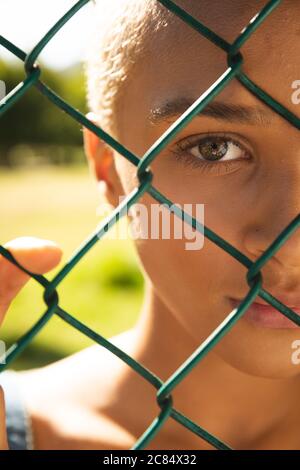Close up portrait of mixed race alternative woman with short blonde hair out and about in the city on a sunny day, looking through a chain link fence. Stock Photo