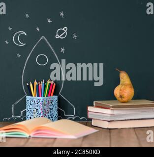 Back to School Concept with Hand Drawn Rocket and stars on Blackboard. Colored pencils and books on a wooden table. Stock Photo