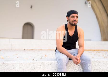Young handsome bearded Indian man thinking while sitting in the streets outdoors Stock Photo