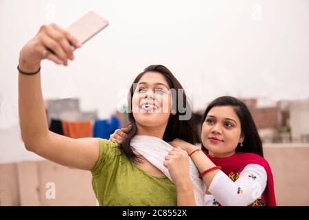 Beautiful Indian young women taking selfie together through smart phone in fresh air and spending their leisure at day time. Stock Photo