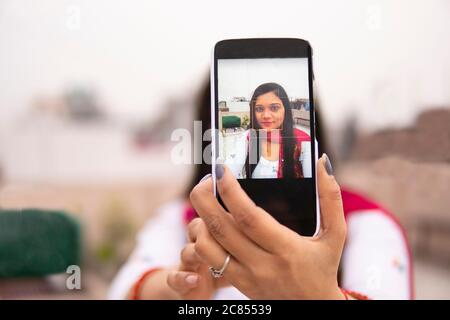Beautiful, happy Indian young woman taking selfie through smartphone and looking at the camera. She is wearing traditional dress salwar Kameez. Stock Photo