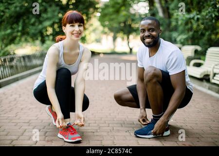 Runners couple, African guy and Caucasian girl, tying running shoes laces at park, smiling and looking at camera. Healthy lifestyle, jogging Stock Photo