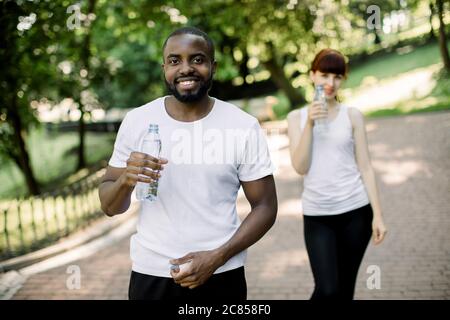 Portrait of handsome joyful smiling African man holding bottle of water, resting after morning workout in city park. His pretty smiling Caucasian Stock Photo