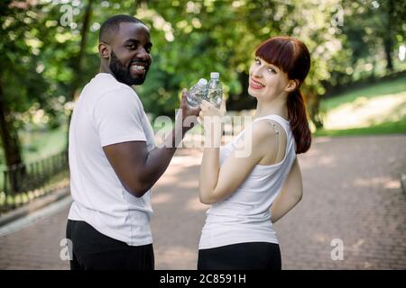 Sport, fitness, jogging and lifestyle concept. Cheerful smiling multiethnical couple with bottles of water, posing to camera during their break after Stock Photo