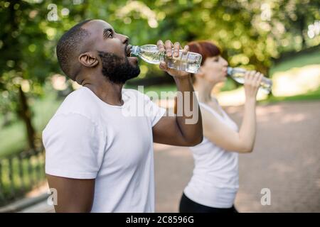 Multiethnical male and female friends drinking water from bottle after fitness sport exercise at city park. Smiling couple with bottles of cold drink Stock Photo
