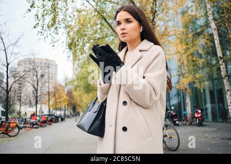 Attractive businesswoman in coat thoughtfully using cellphone waiting on city street Stock Photo