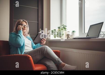 Busy senior woman with blonde hair and eyeglasses holding a tablet and talking on phone while working from home Stock Photo