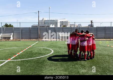 Multi ethnic team of male five a side football players wearing a team strip training at a sports field in the sun, standing in huddle moticativating e Stock Photo