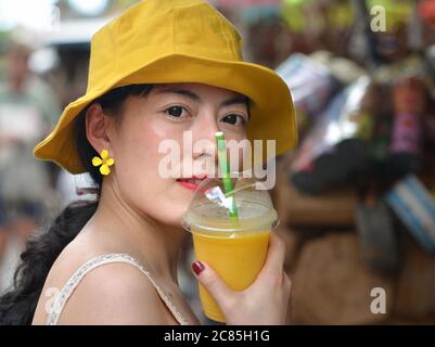 Young Asian woman with yellow floppy hat wears dark contact lenses and holds a plastic cup with yellow fruit juice. Stock Photo