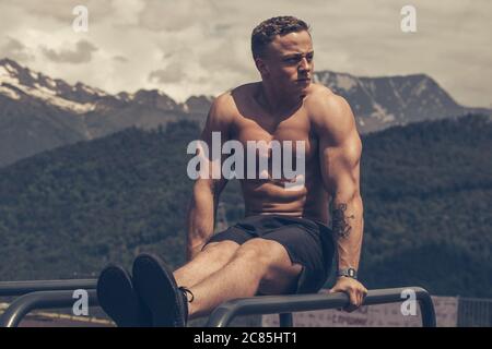 young blonde male athlete with focused and tensed face expression exercising in the park does press ups on parallel bars over mountains background . Stock Photo