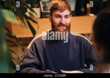Young cheerful bearded man happily looking away on meeting in cozy cafe Stock Photo