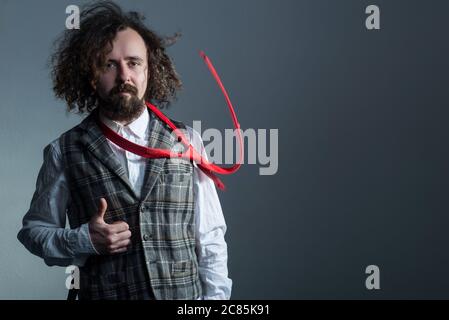 studio portrait in motion of a young bearded, curly haired guy of thirty years, in a plaid vest, and a flying red tie, waving long hair, holds his thu Stock Photo