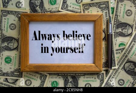 Wooden picture frame with inscription 'always believe in yourself' on beautiful background from dollar bills and metalic pen. Concept. Stock Photo