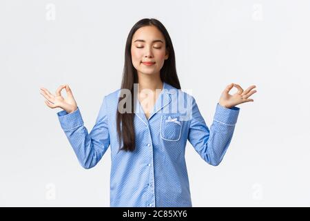 Calm and happy smiling asian girl in blue pajamas close eyes, meditating before sleep or in morning, looking relieved and peaceful, practice yoga Stock Photo