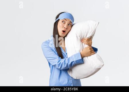 Surprised asian girl in awe wearing sleeping mask and pajamas as lying in bed and hugging pillow, looking startled upper left corner, drop jaw and say Stock Photo