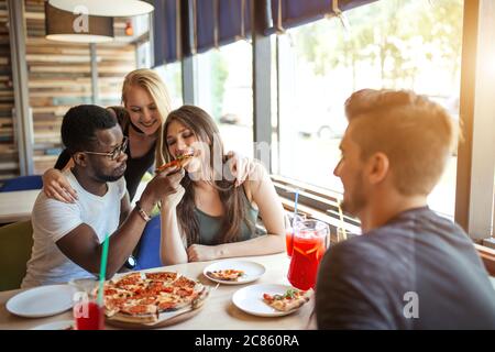 Cheerful happy male and female students treating each other with organic pizza with different toppings and non alchoholic cocktails, having fun and en Stock Photo